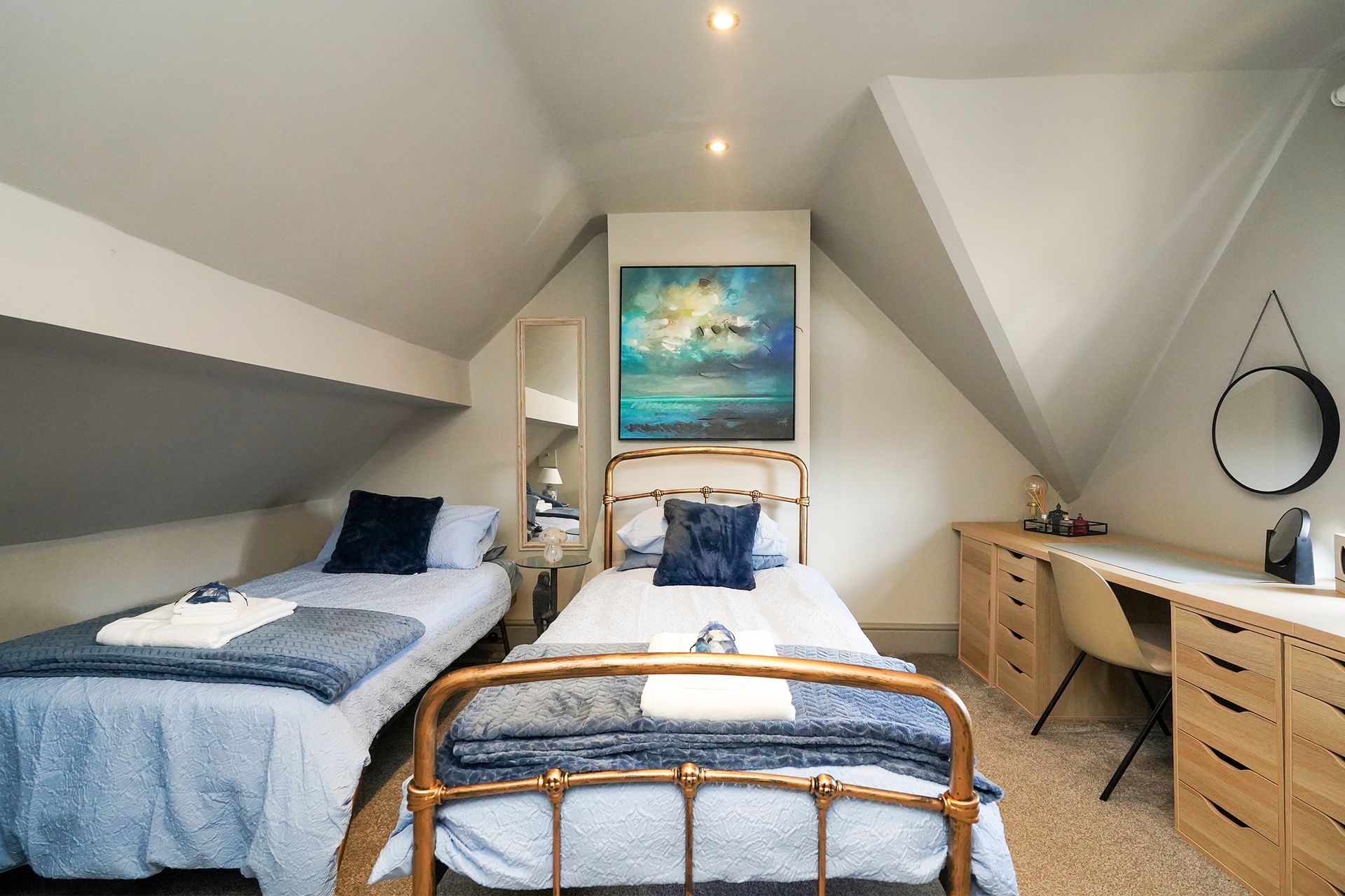 Small attic bedroom with two twin beds, a desk, and a round mirror under slanted white ceilings.