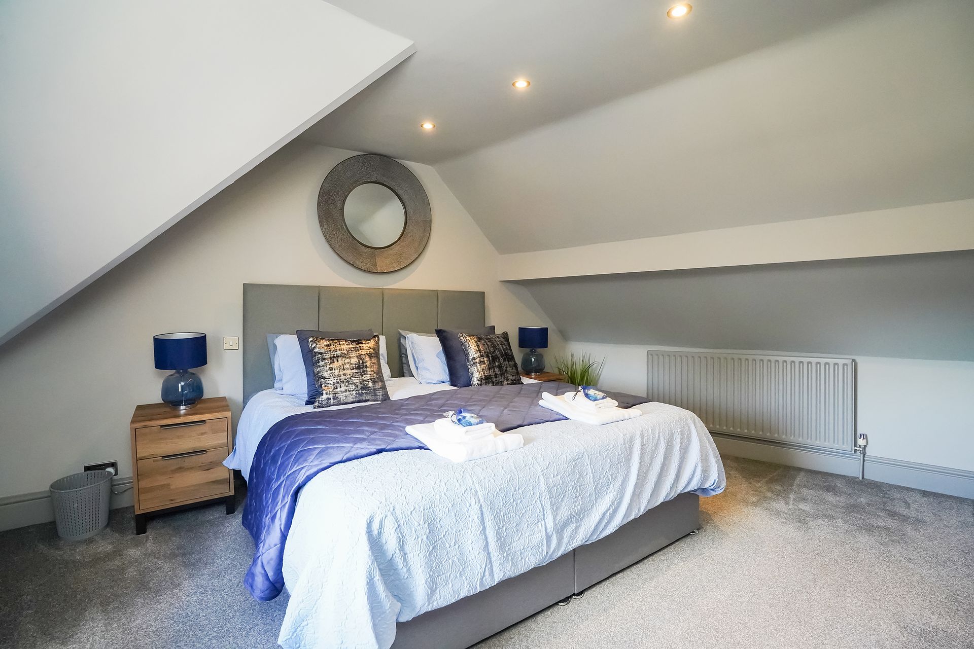 Attic bedroom with a large bed, blue accents, bedside table, mirror, and radiator under sloped ceilings