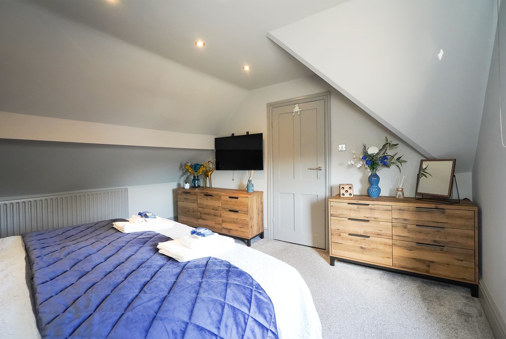 Bright attic bedroom with blue bedspread, two wooden dressers, and a white door under a sloped ceiling