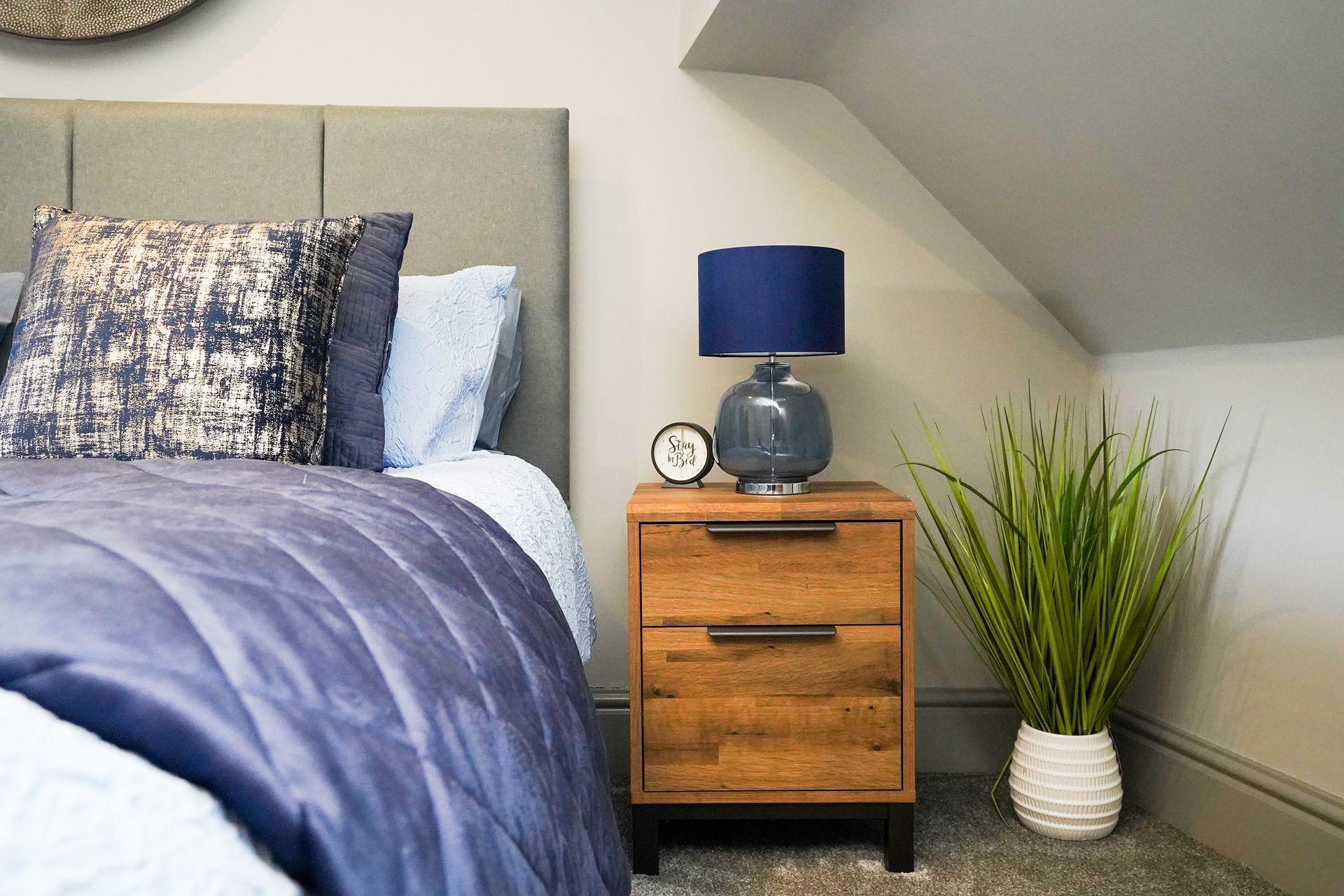 Modern bedroom corner with bed, blue lamp on wooden nightstand, and potted plant