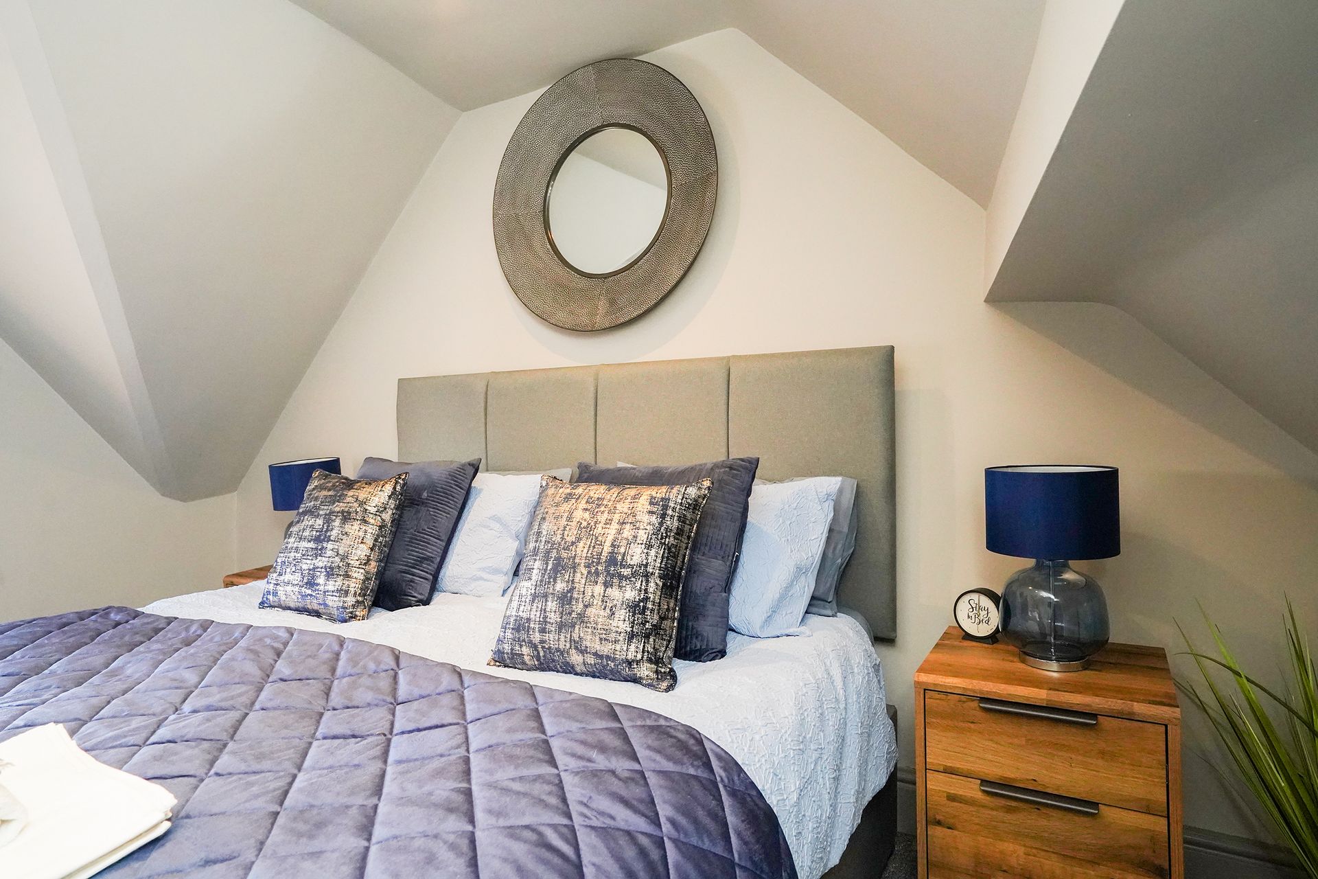 Cozy attic bedroom with a gray bed, patterned pillows, and a wooden nightstand with a blue lamp.