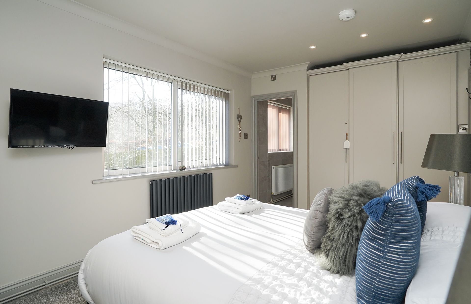 Bright bedroom with a white bed, wall-mounted TV, window blinds, and built-in wardrobe cabinets.