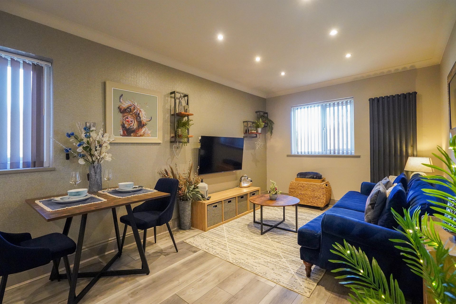 Cozy living room with blue sofas, dining table, TV, and large windows with blinds