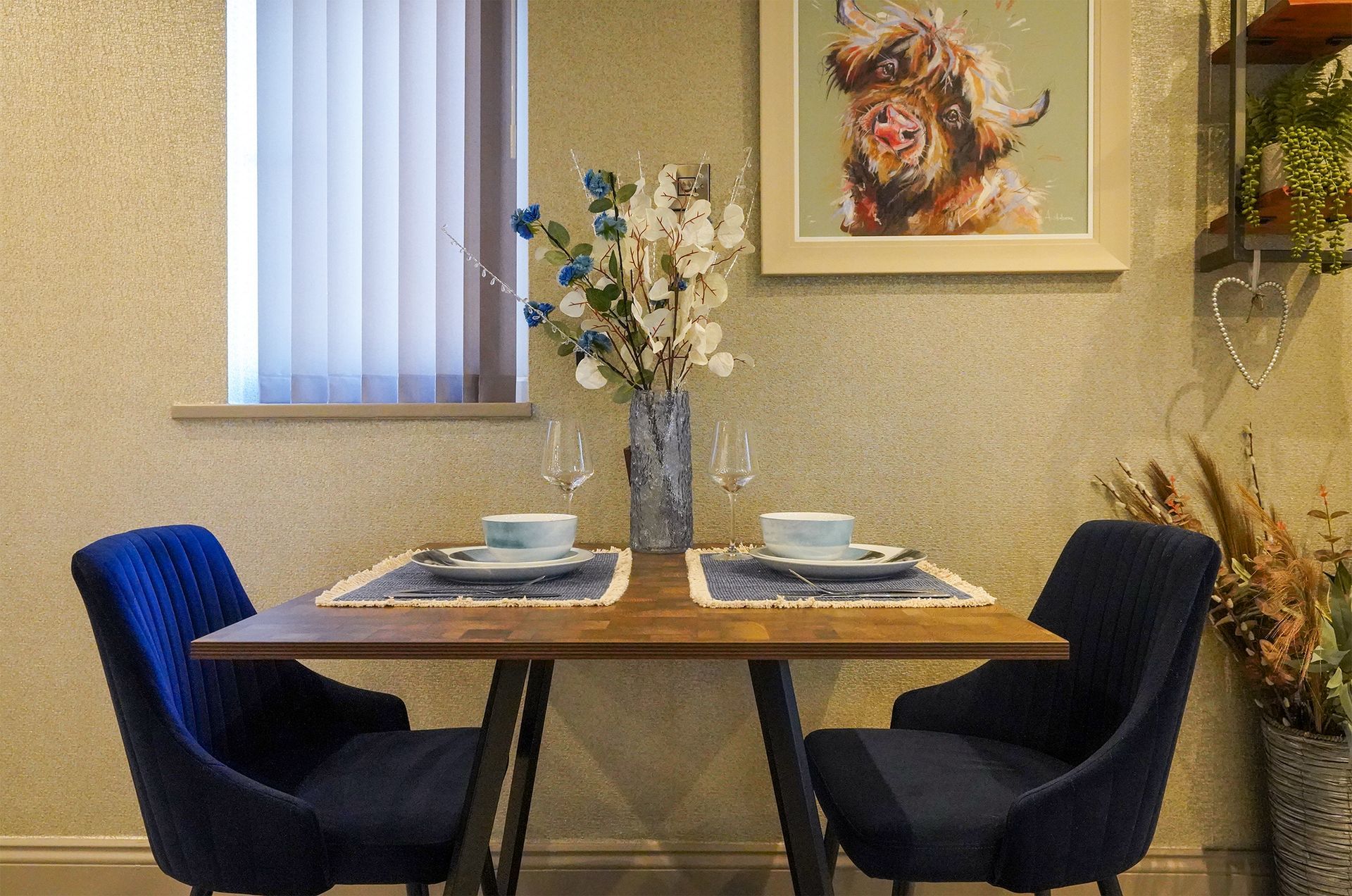 Modern dining nook with two blue chairs, set table, floral centerpiece, and wall art