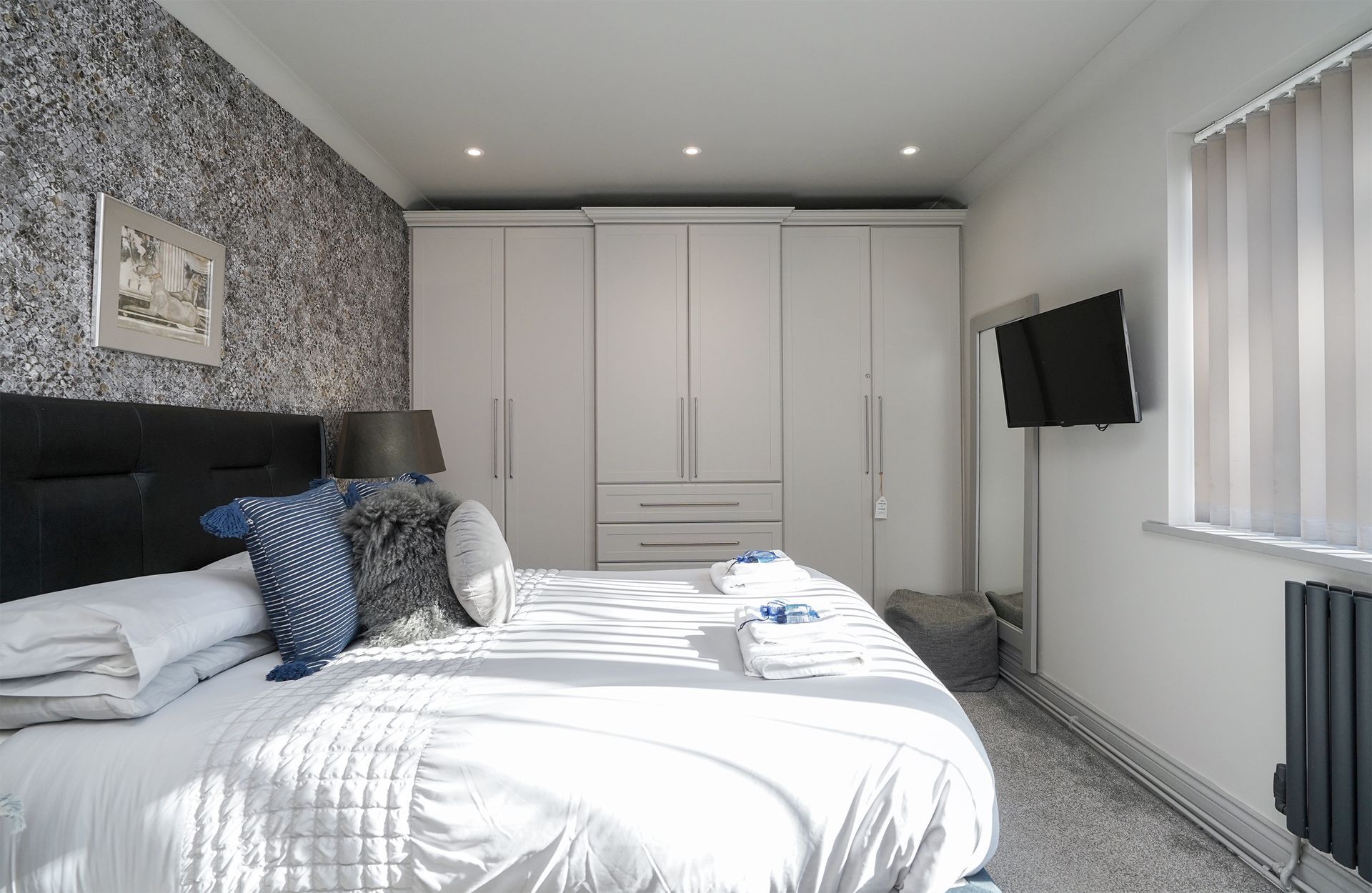 Bright modern bedroom with a bed, wardrobe, wall-mounted TV, and window blinds