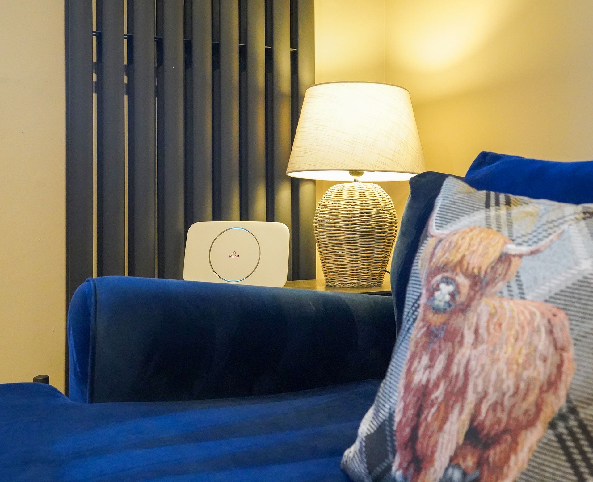 Blue sofa with a patterned buffalo pillow beside a lit table lamp and vertical blinds in a warm room