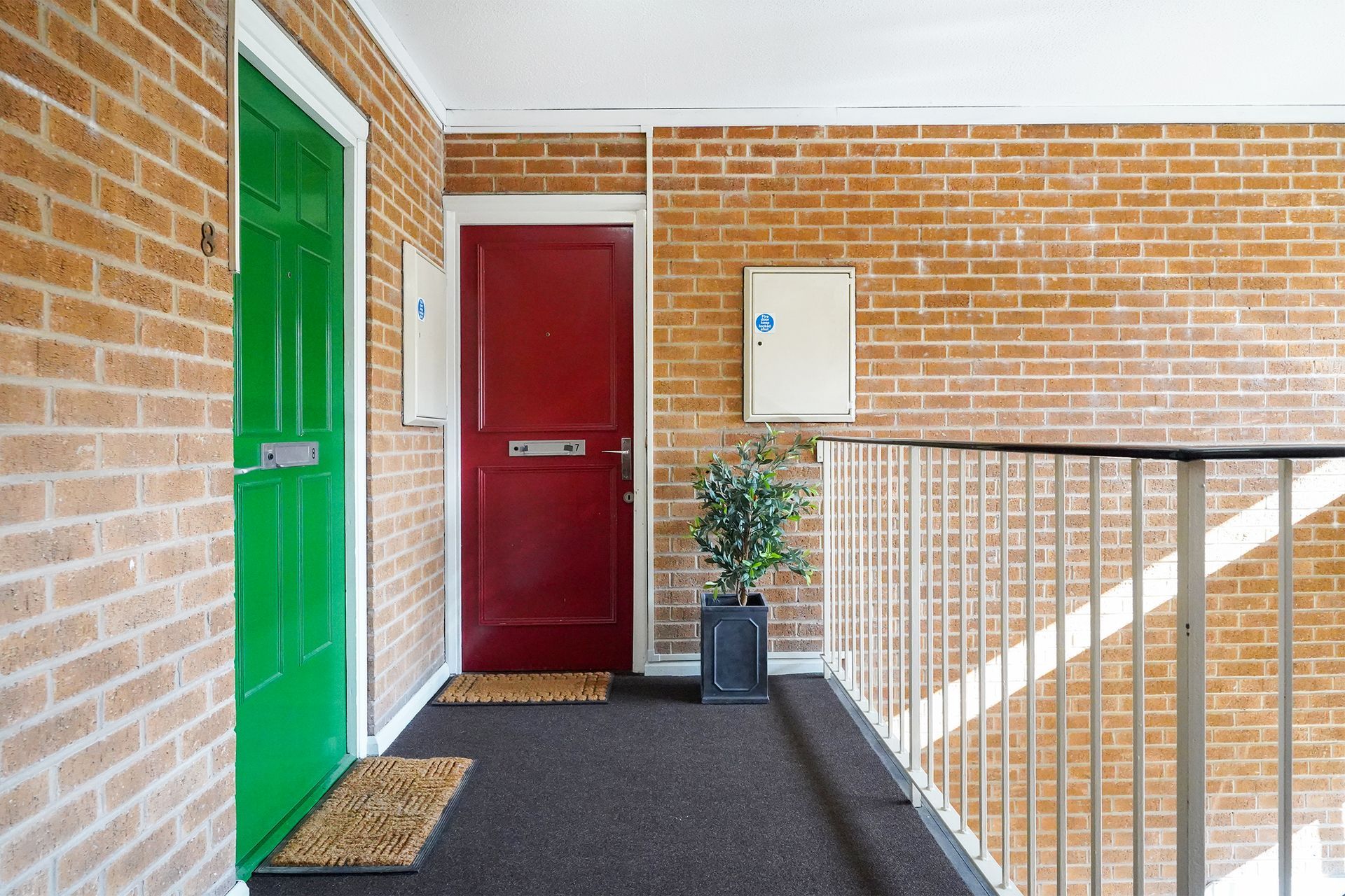 Apartment corridor with green and red doors, brick walls, black floor, and a potted plant near a railing