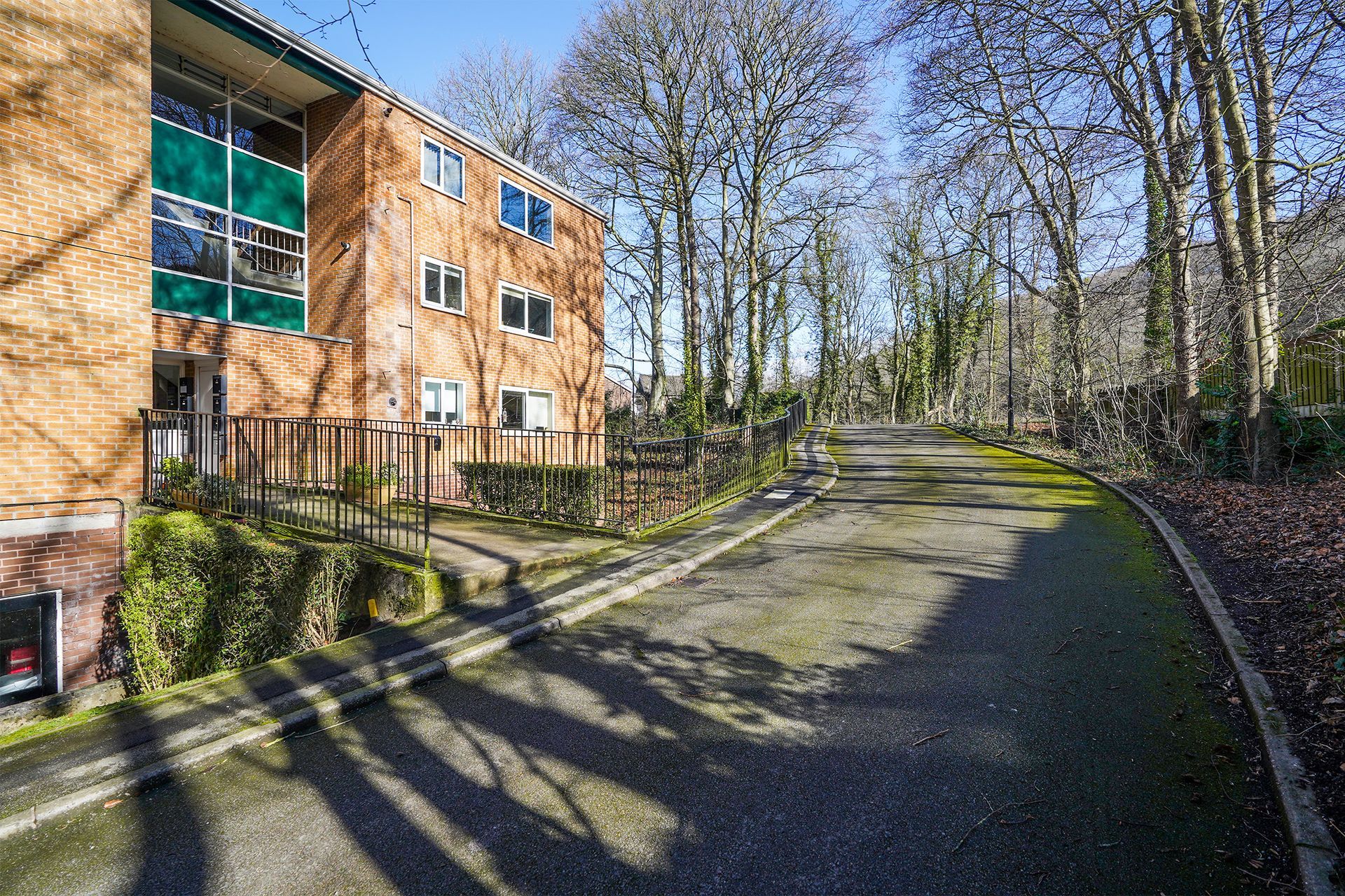 Brick apartment building beside a curving paved path, with trees and bright sunlight casting shadows.