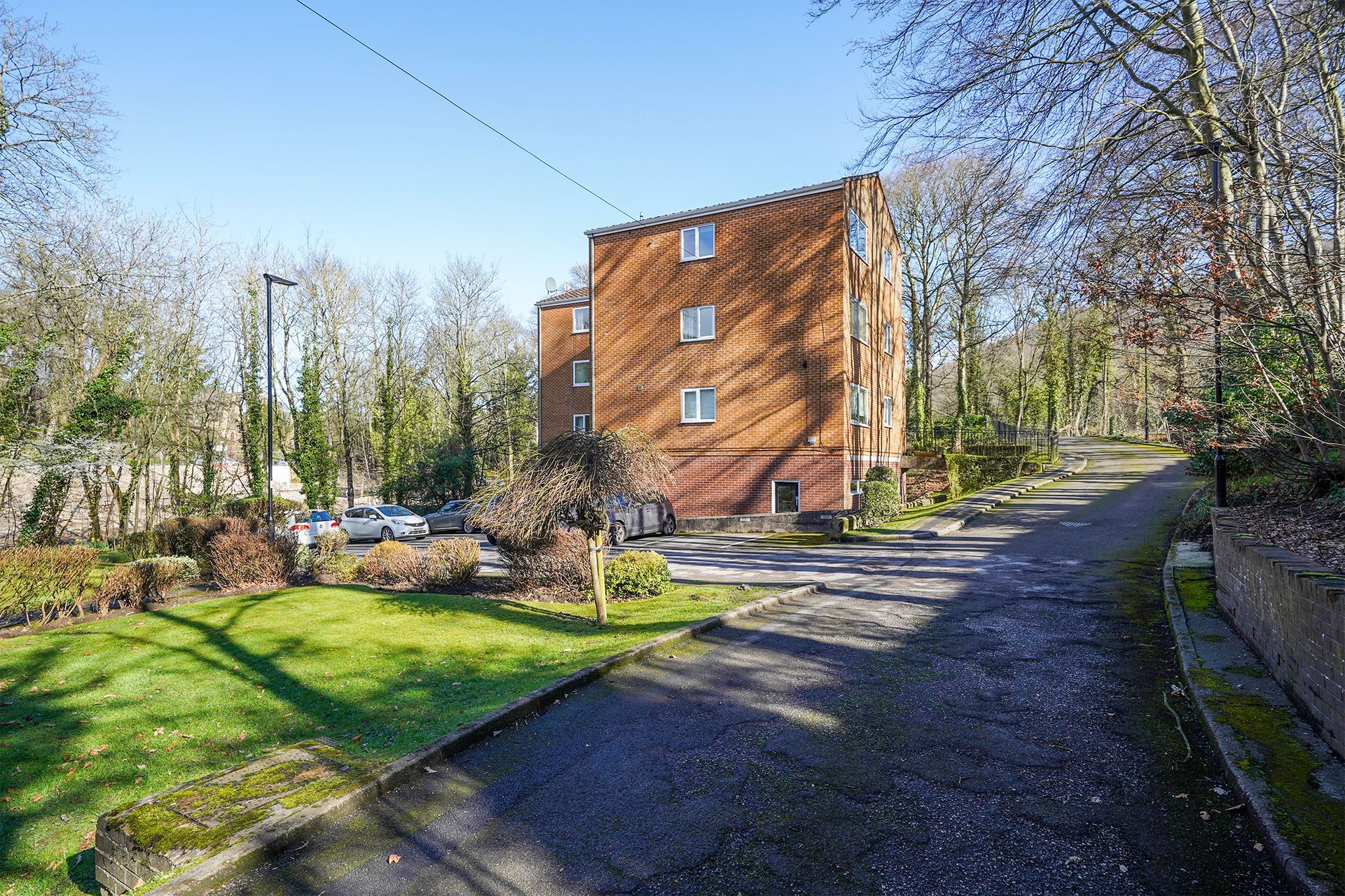 Brick apartment building beside a gravel path and green lawn in a sunny wooded area