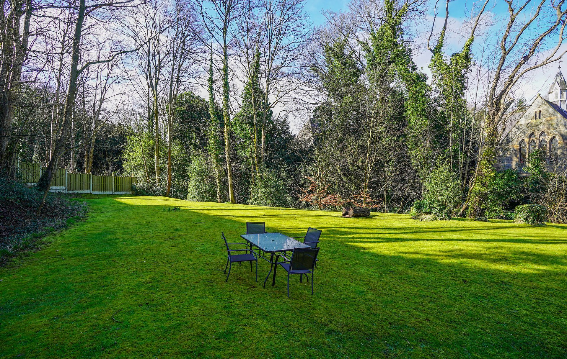 Green park lawn with a small table and two chairs near a tree-lined path under bright sunlight