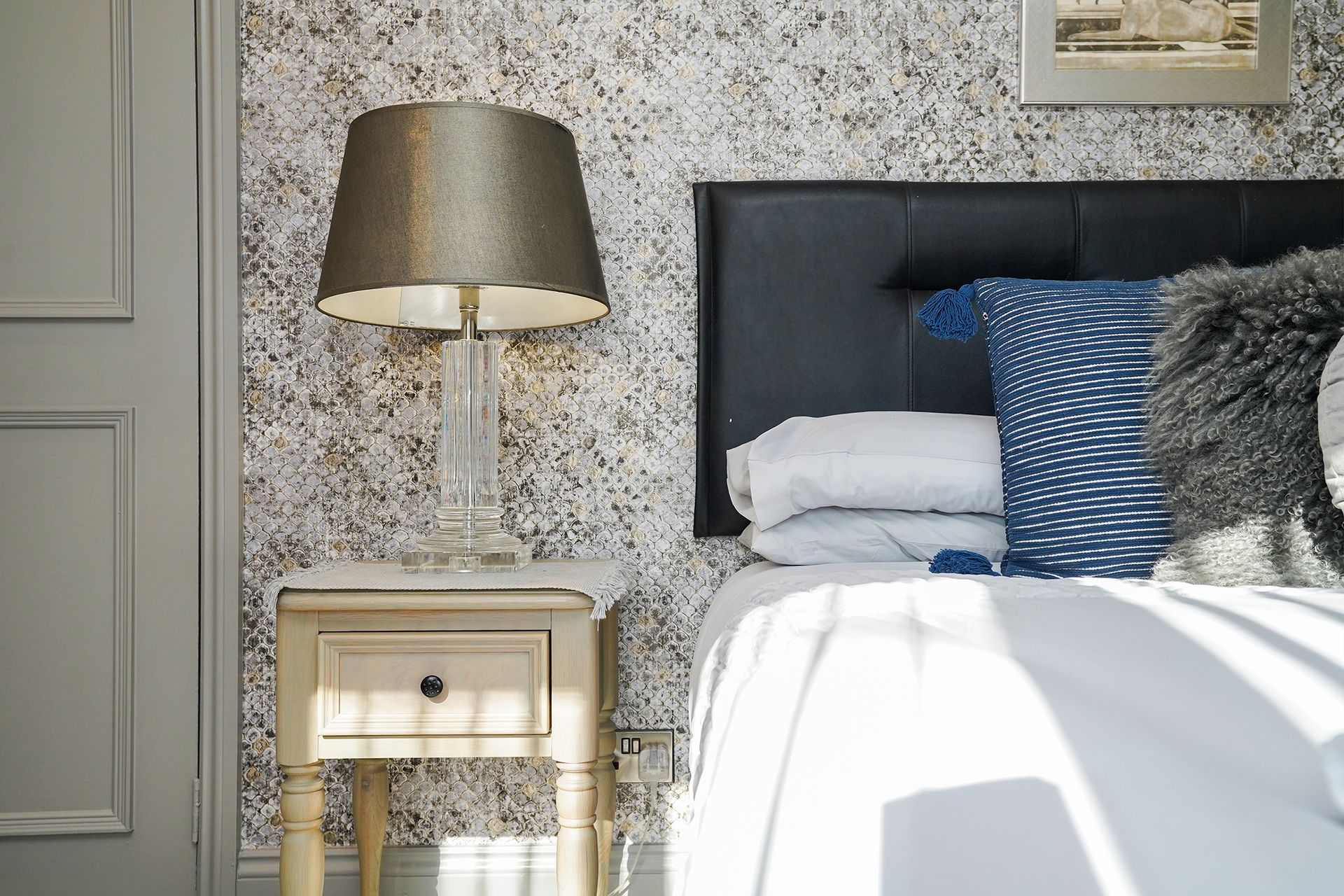Bedroom with a dark headboard, blue and gray pillows, bedside lamp, and floral wallpaper