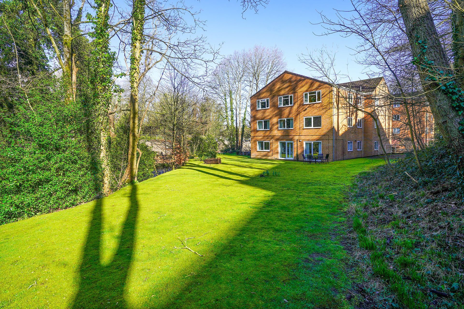 Sunny grassy yard beside a brick apartment building with trees and long shadows