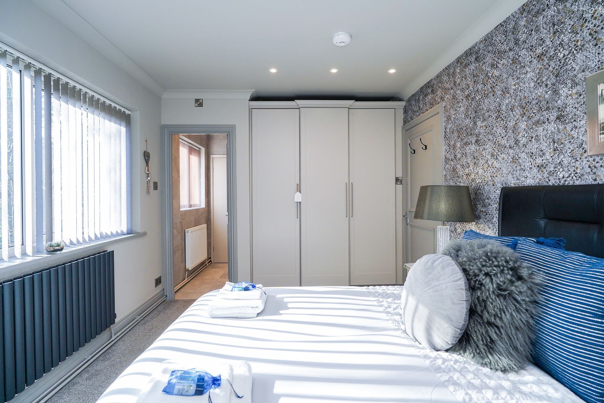 Bright bedroom with white bed, blue accents, wardrobe, window blinds, and patterned wallpaper.