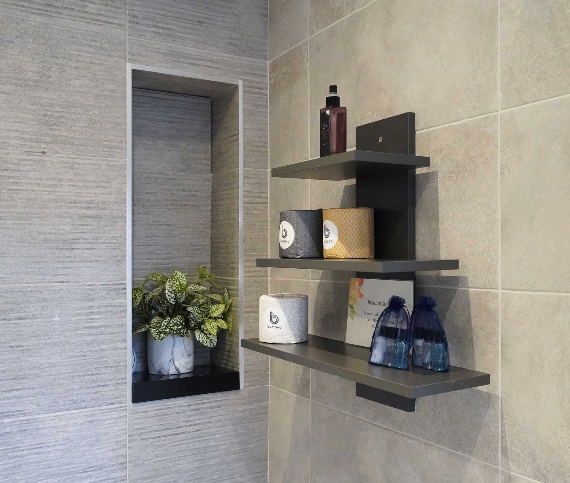 Modern bathroom wall with dark shelves holding toiletries, decor, and blue bottles beside a recessed plant niche