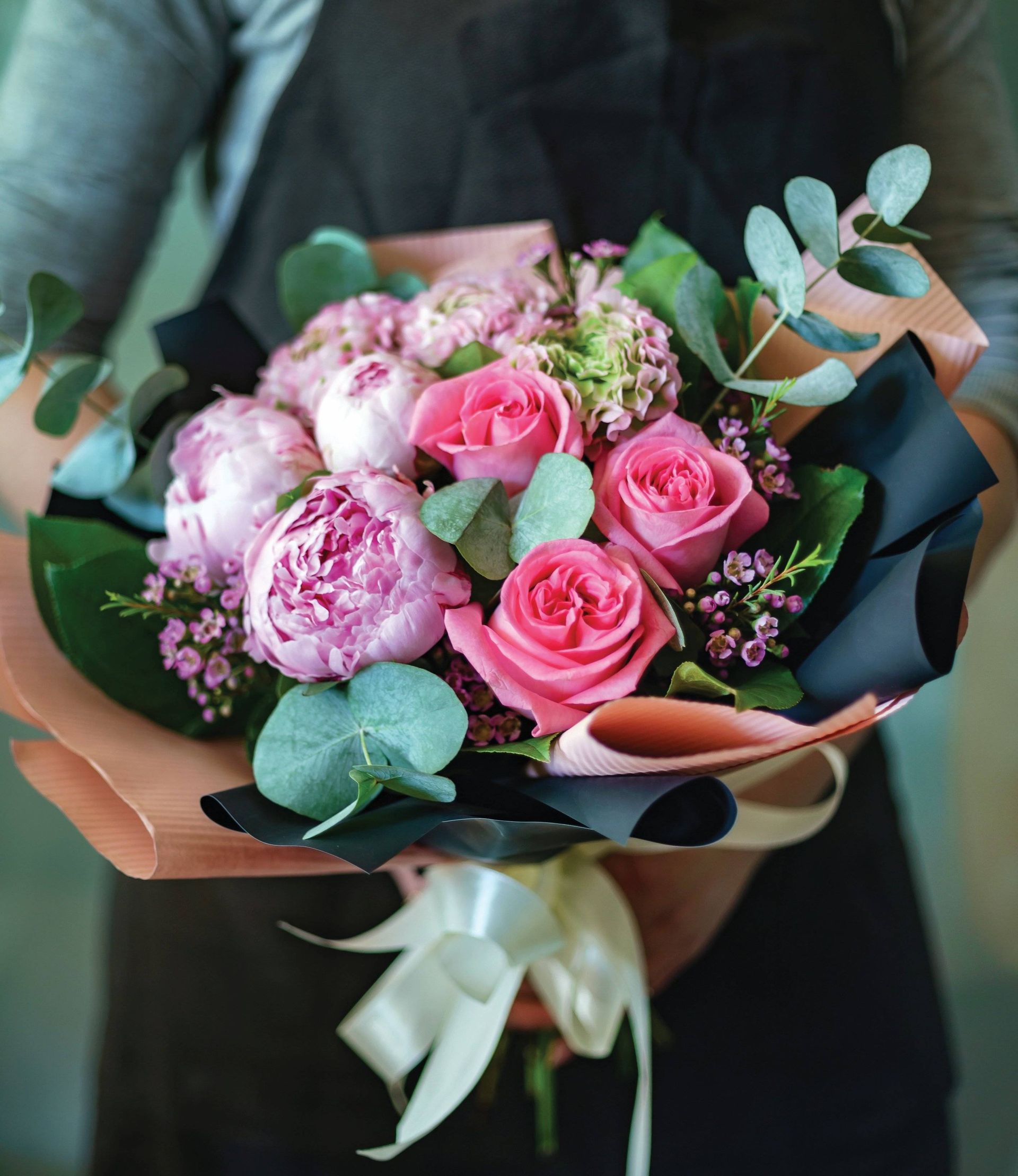 A bouquet of pink roses and peonies wrapped in dark paper with a ribbon