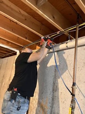 Man using a drill to install wiring overhead in an unfinished basement with exposed beams and concrete walls