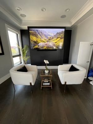 Modern living room with dark walls, two white chairs, a glass table, and a large landscape TV display.