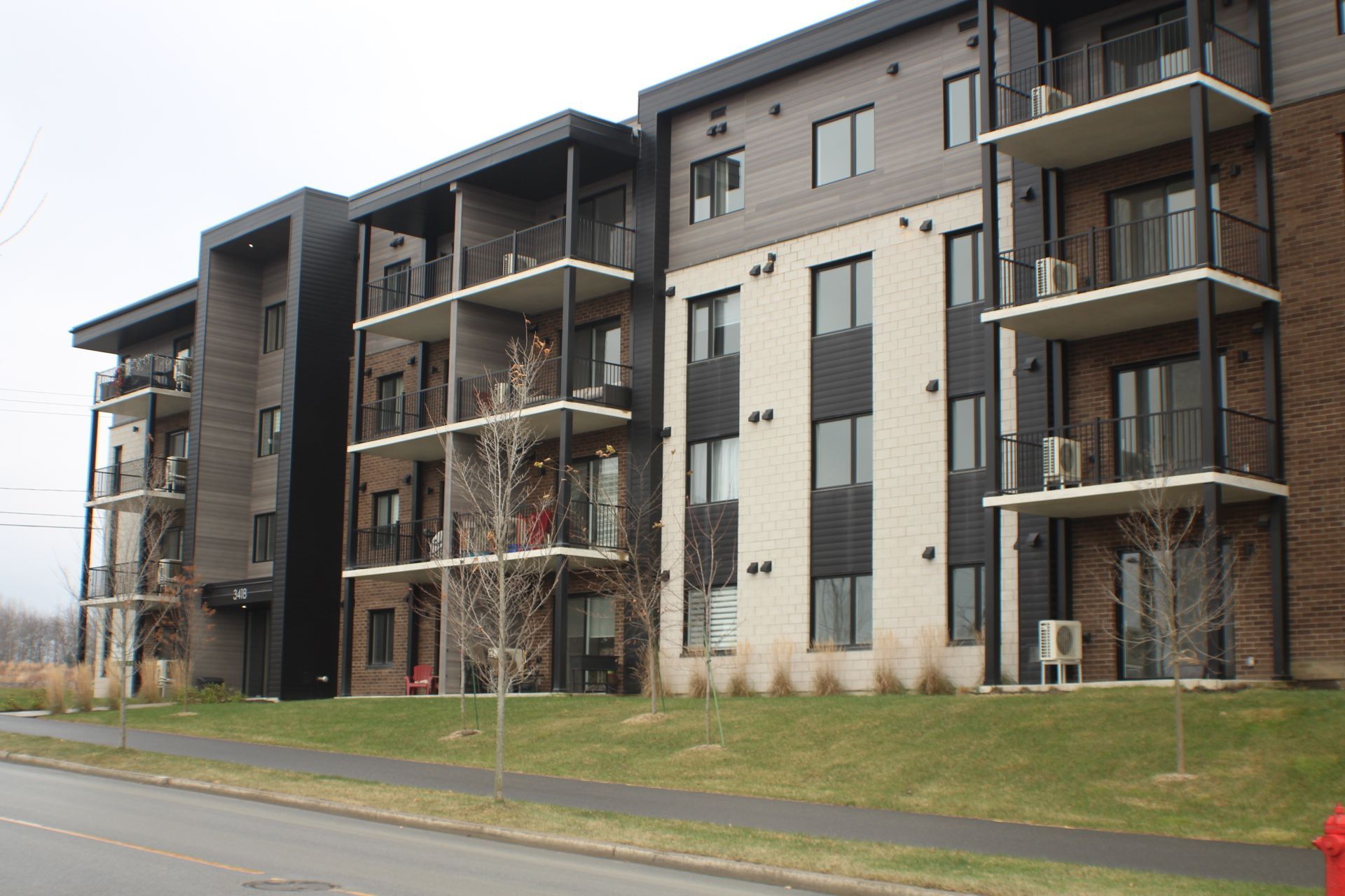 Immeuble d'appartements à plusieurs étages avec balcons; façade en brique et couleur claire, pelouse herbeuse, ciel couvert.