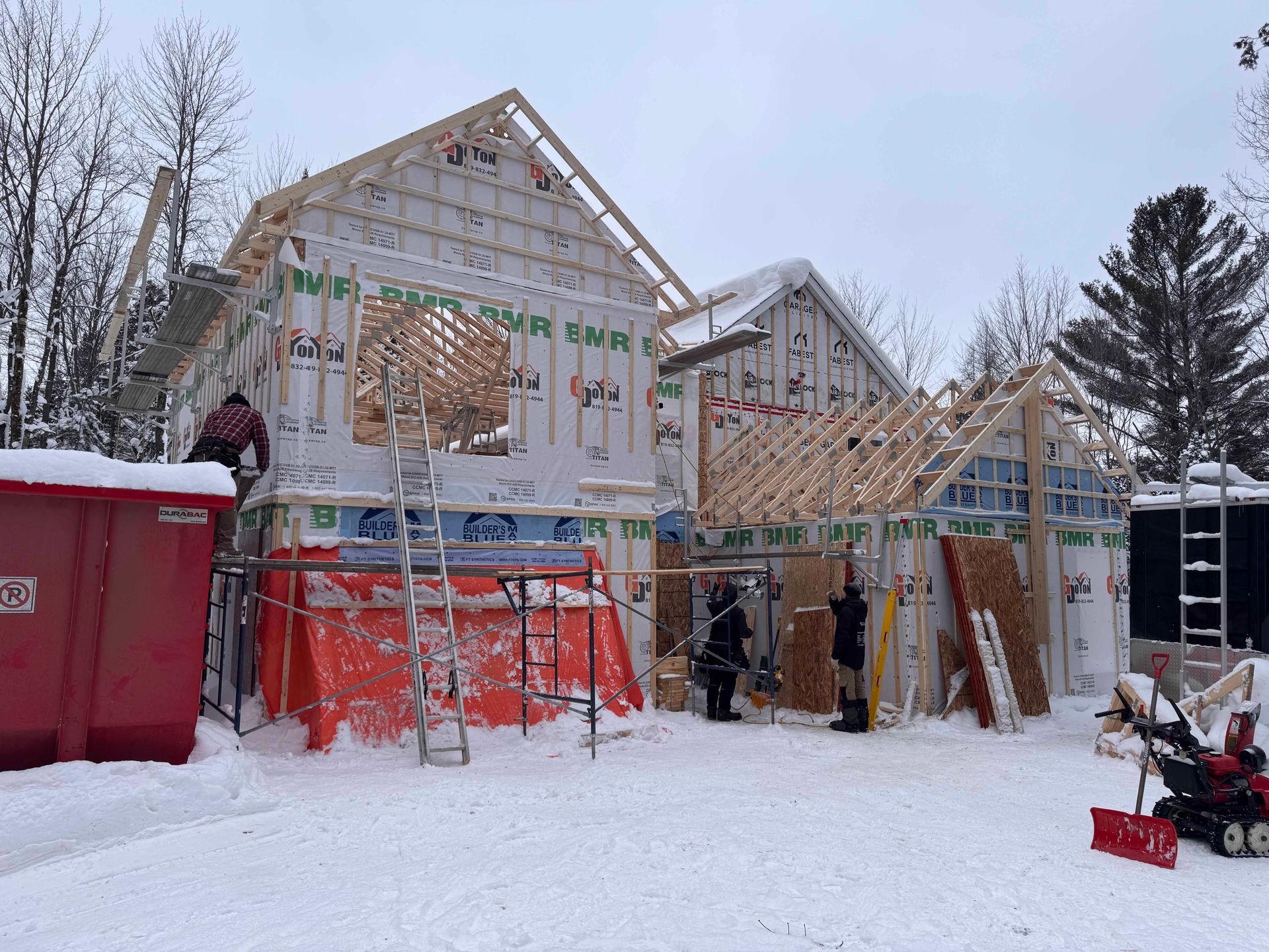 Chantier de construction, ossature de maison en construction dans un décor extérieur enneigé, ouvriers présents.