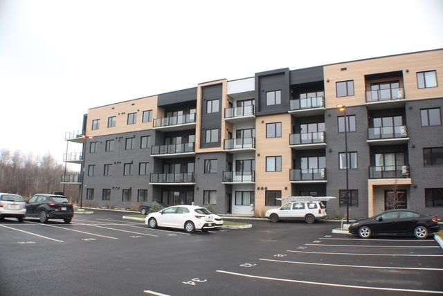 Immeuble d'appartements moderne avec des voitures garées sur un terrain pavé sous un ciel couvert.