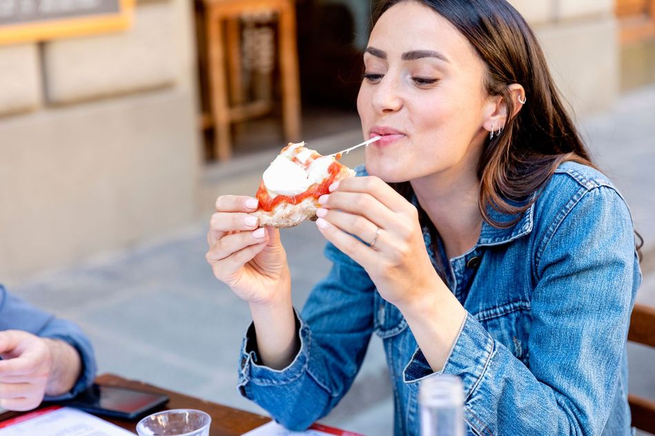 Donna che mangia la pizza all'aperto, tirando fuori il formaggio, sorridendo.