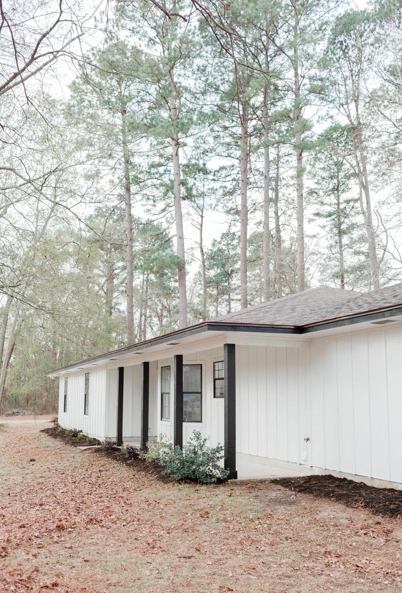 White single-story house with black shutters in a wooded yard, viewed from the front corner.