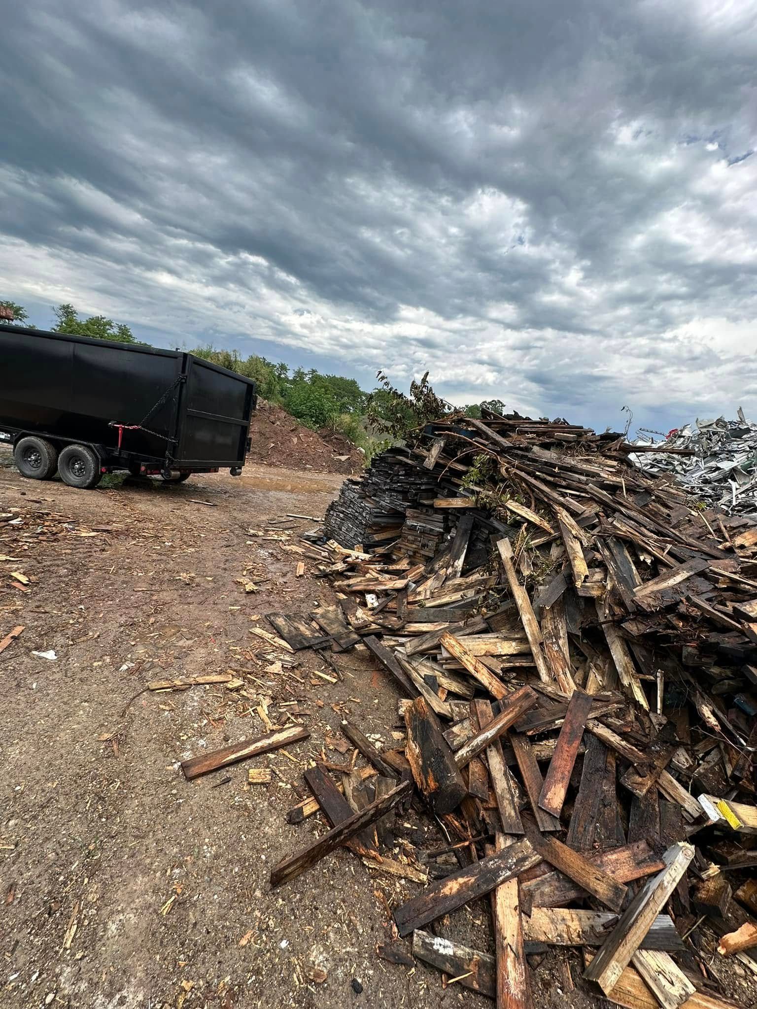 Piles of scrap wood beside a black trailer on a gravel lot under a cloudy sky