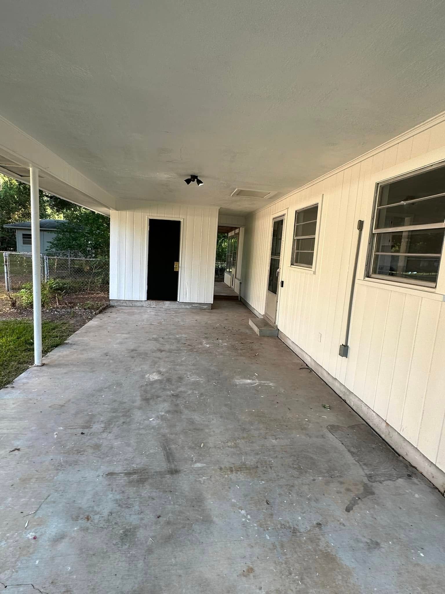 Covered concrete carport beside a cream house with windows and a dark doorway