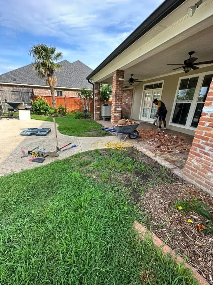 Backyard patio under construction with brick house, tools, wheelbarrow, and worker near glass doors