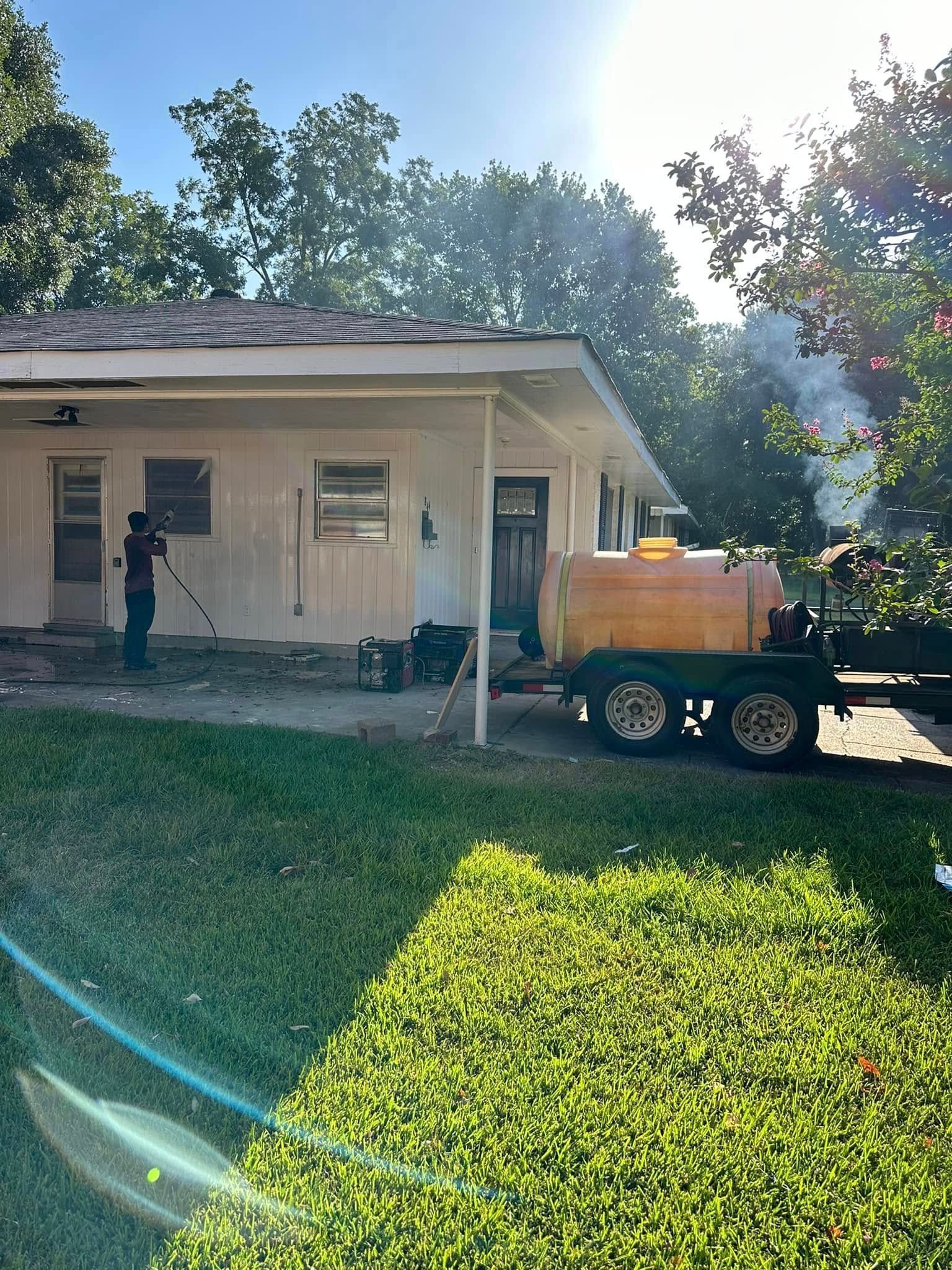 A person pressure washes a house exterior beside a trailer-mounted water tank in bright sunlight.