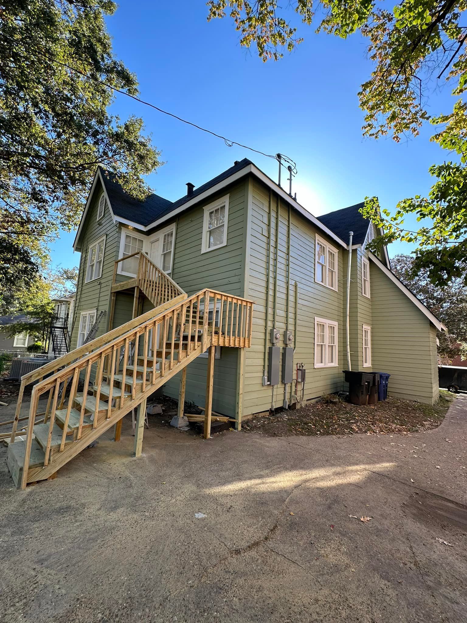 Green two-story house with exterior wooden stairs and a gravel driveway under a clear blue sky