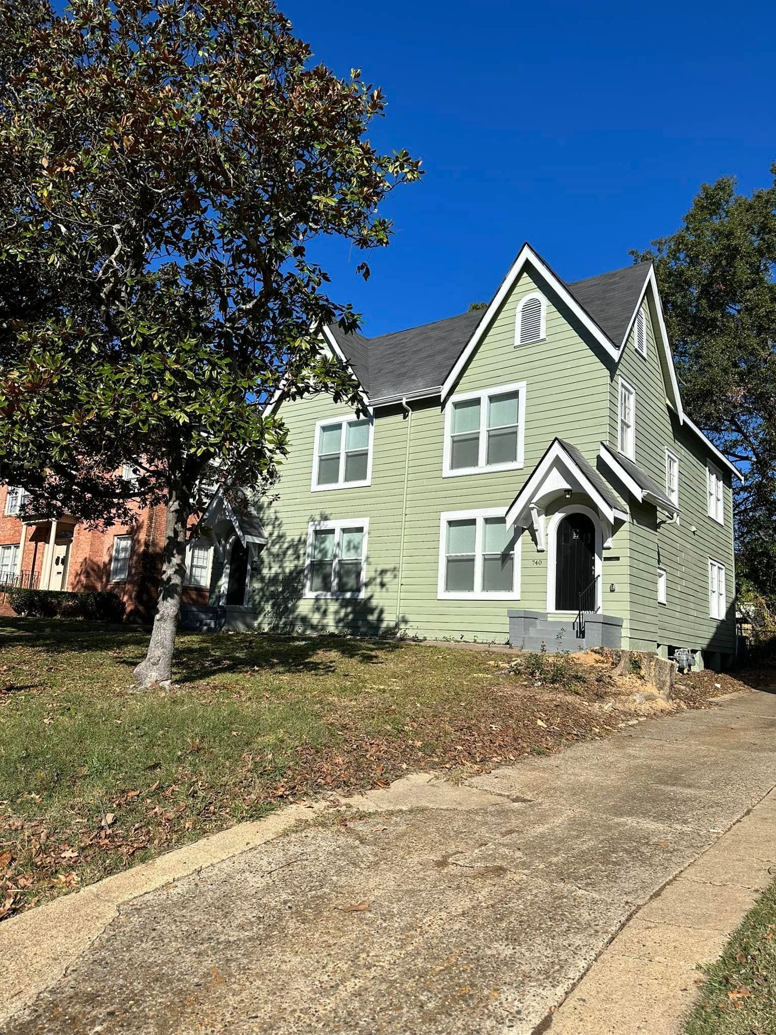 Light green house with steep gables and a front porch, beside a tree and sloped driveway under a blue sky