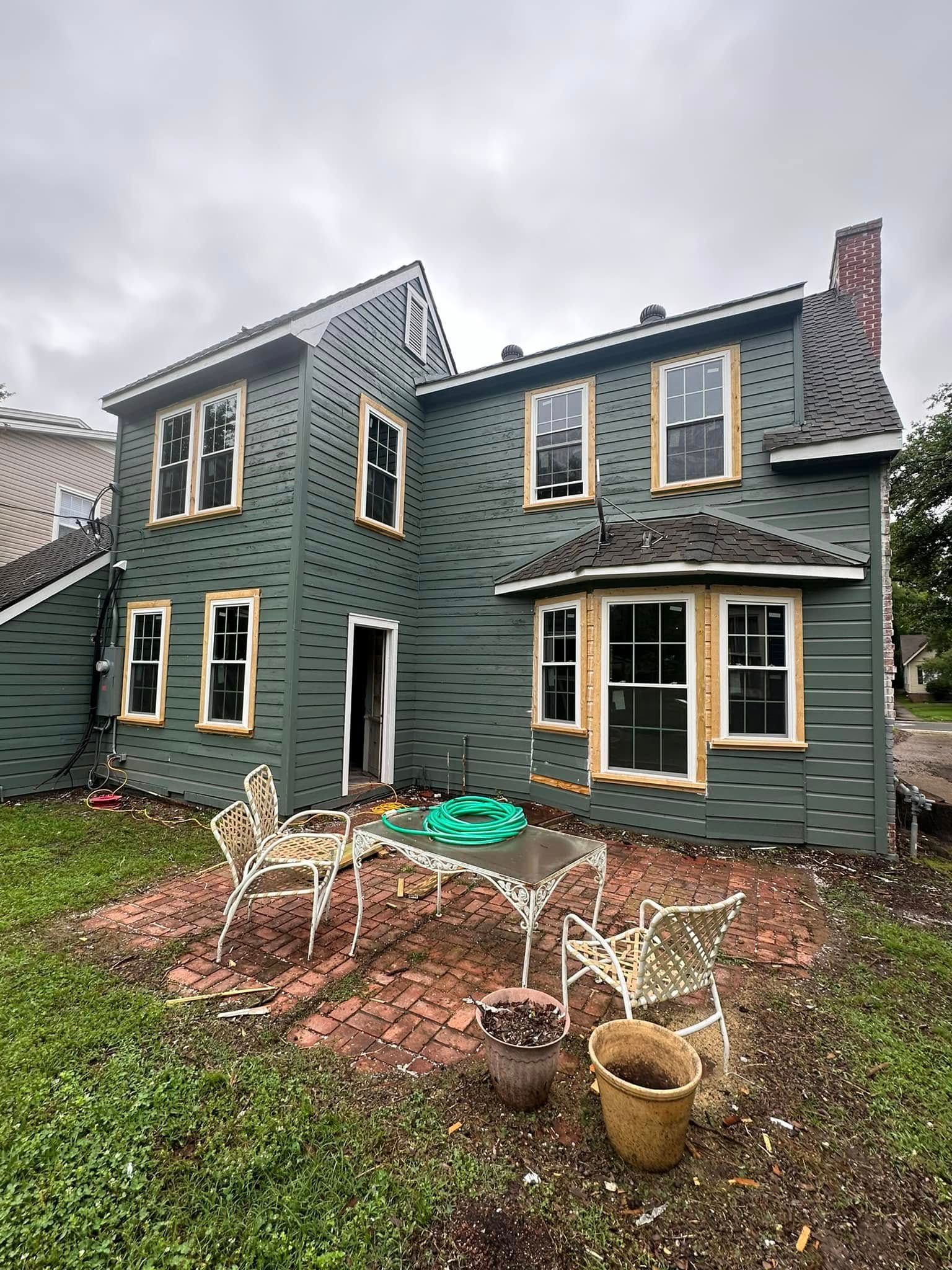 Backyard view of a green two-story house with patio furniture and potted plants on a brick patio.