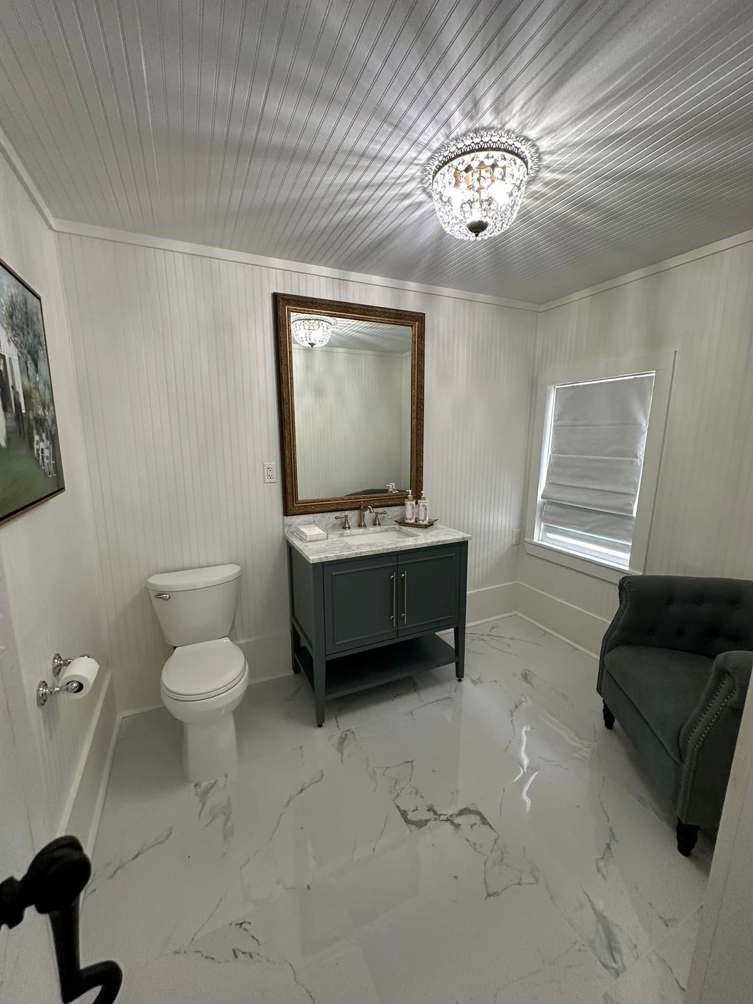 Small bathroom with white marble floor, gray vanity, mirror, toilet, window, and dark chair.