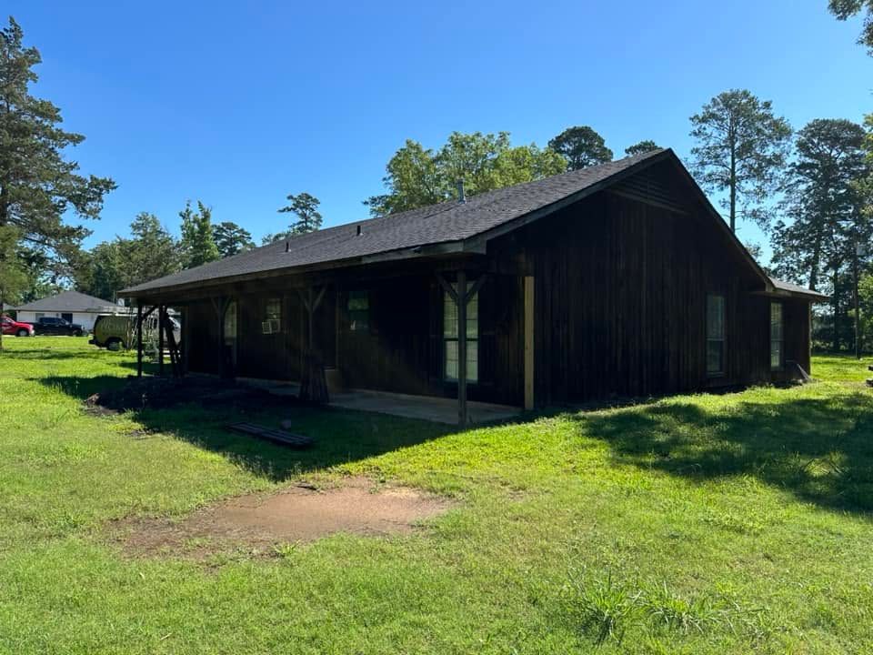 Rustic dark-wood cabin with a porch in a sunny grassy clearing surrounded by trees