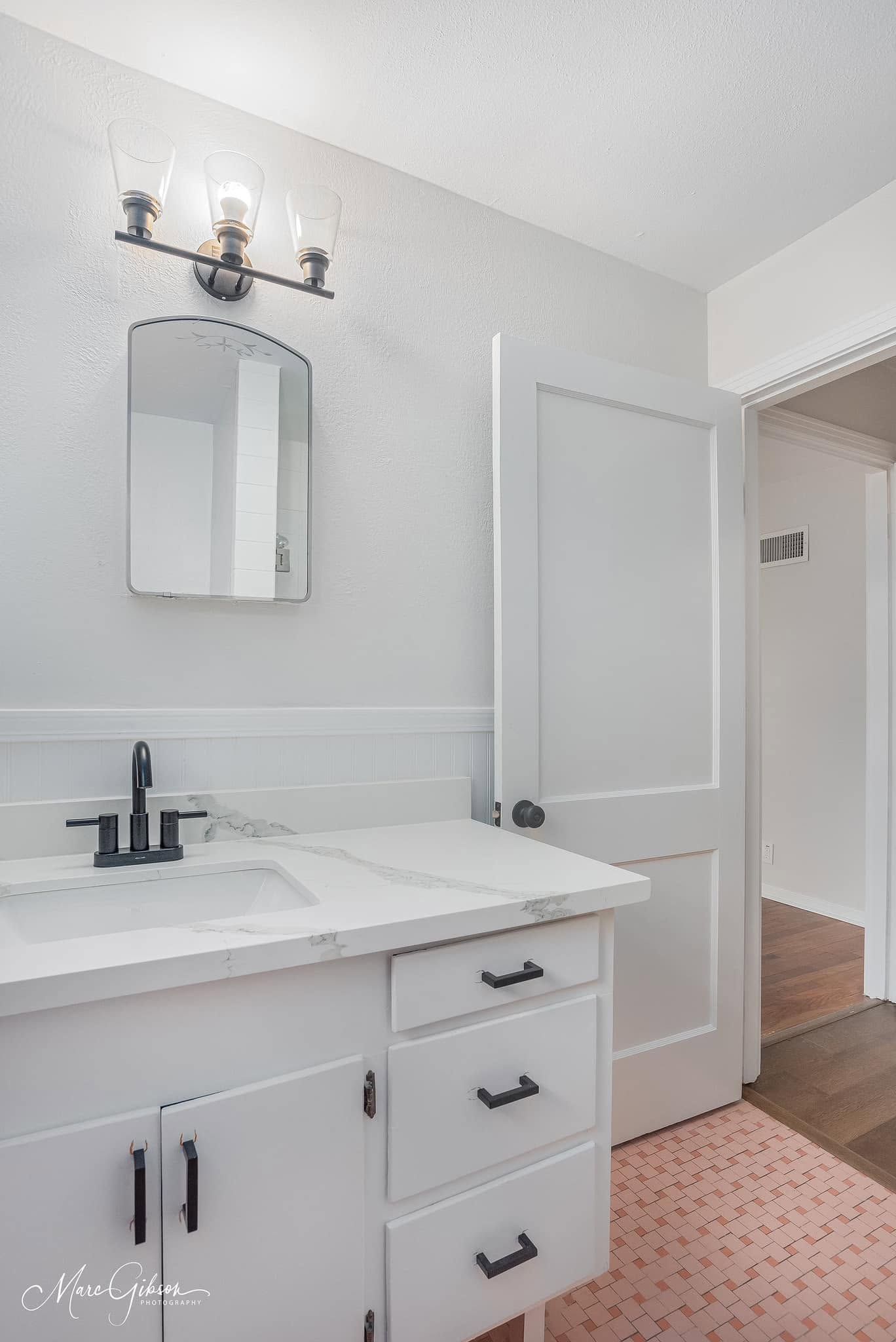 Bright white bathroom vanity with mirror, black faucet, wall sconce, and pink patterned rug