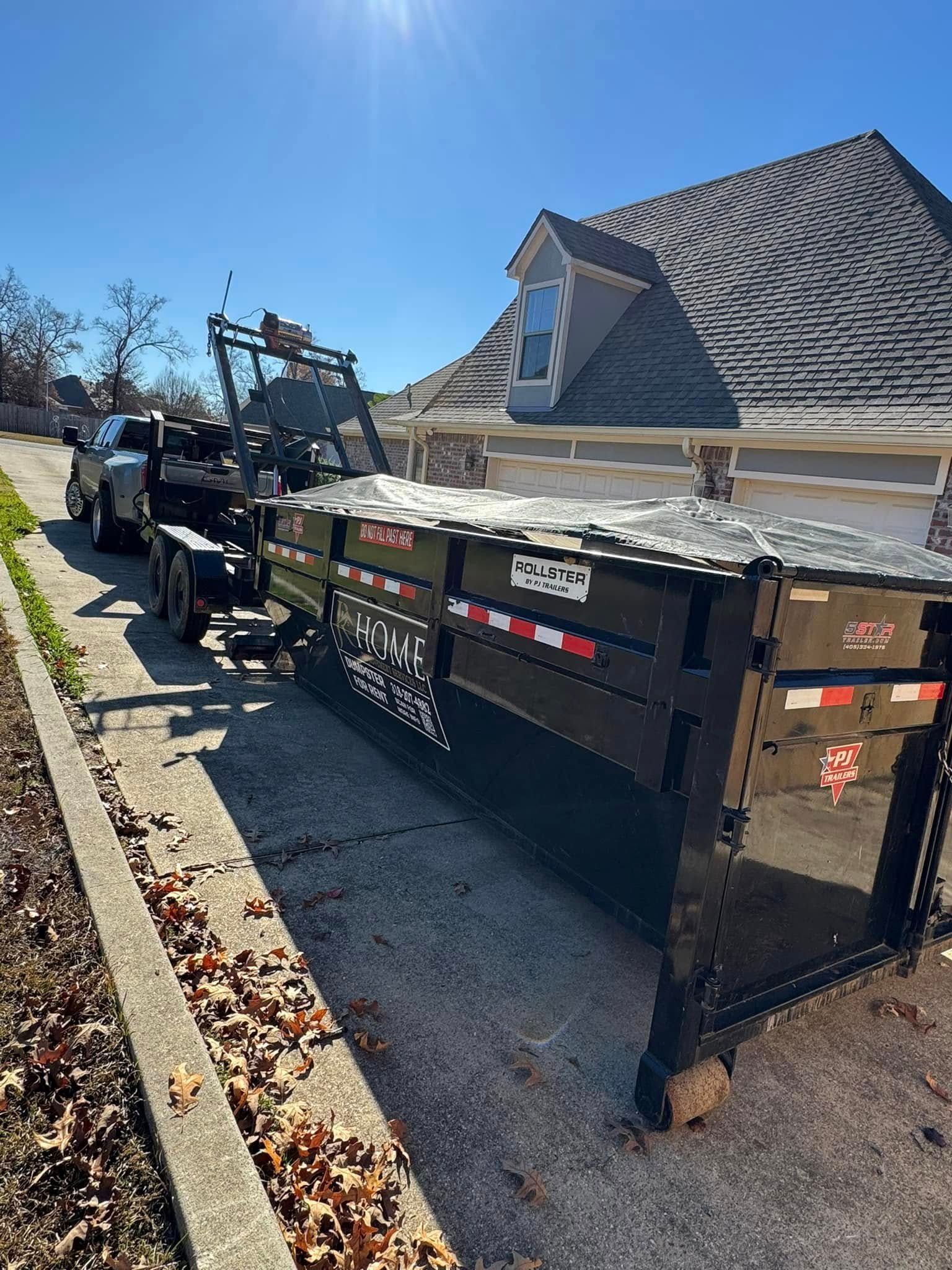 Black dump trailer parked on a residential street beside a house, with a truck and lift equipment attached.