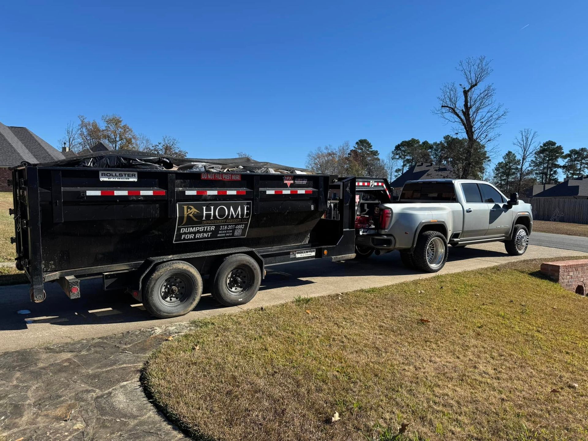 Silver pickup truck towing a black dump trailer on a suburban driveway under a clear blue sky