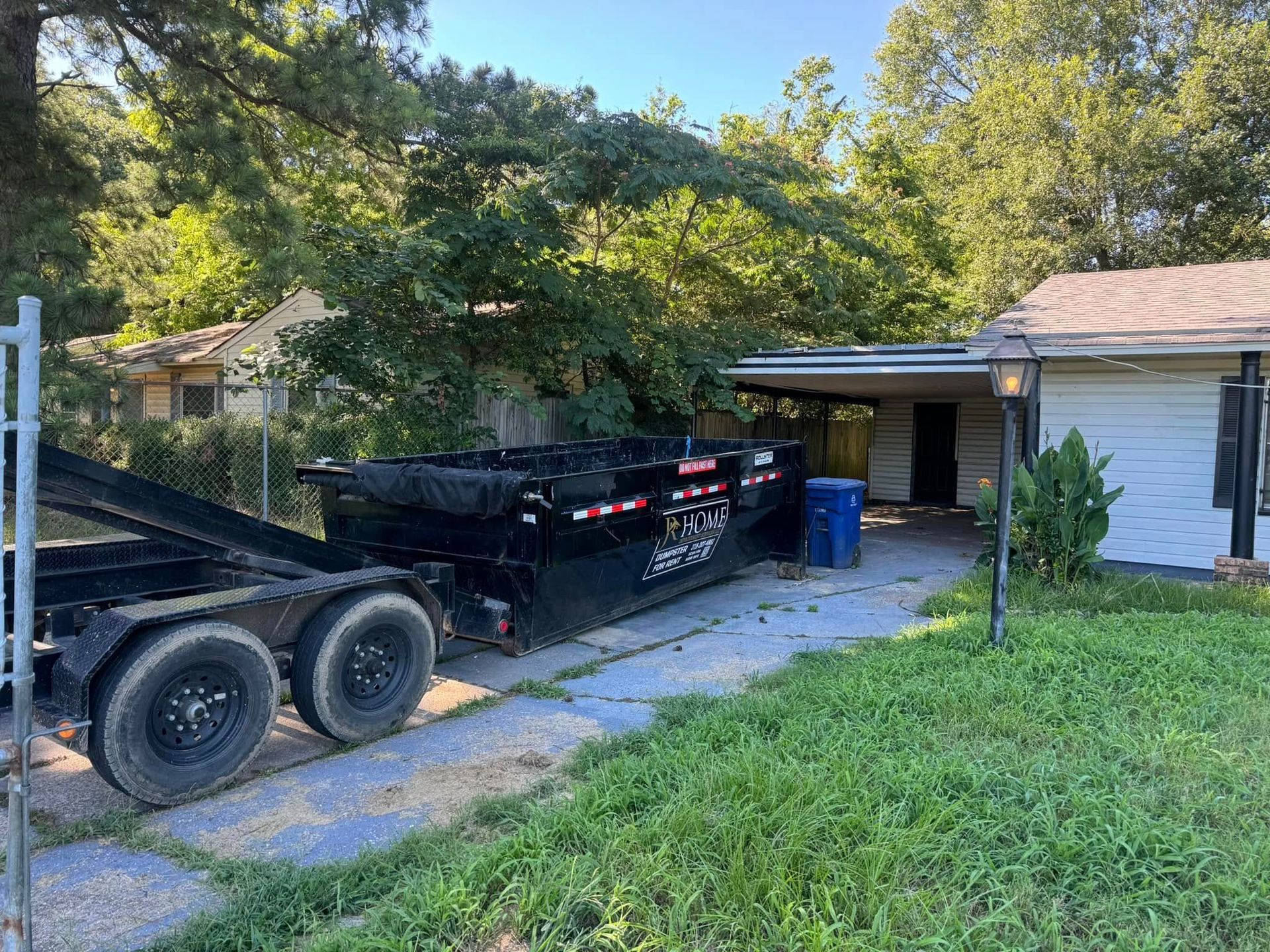 Black dump trailer parked in a driveway beside a white house and green yard