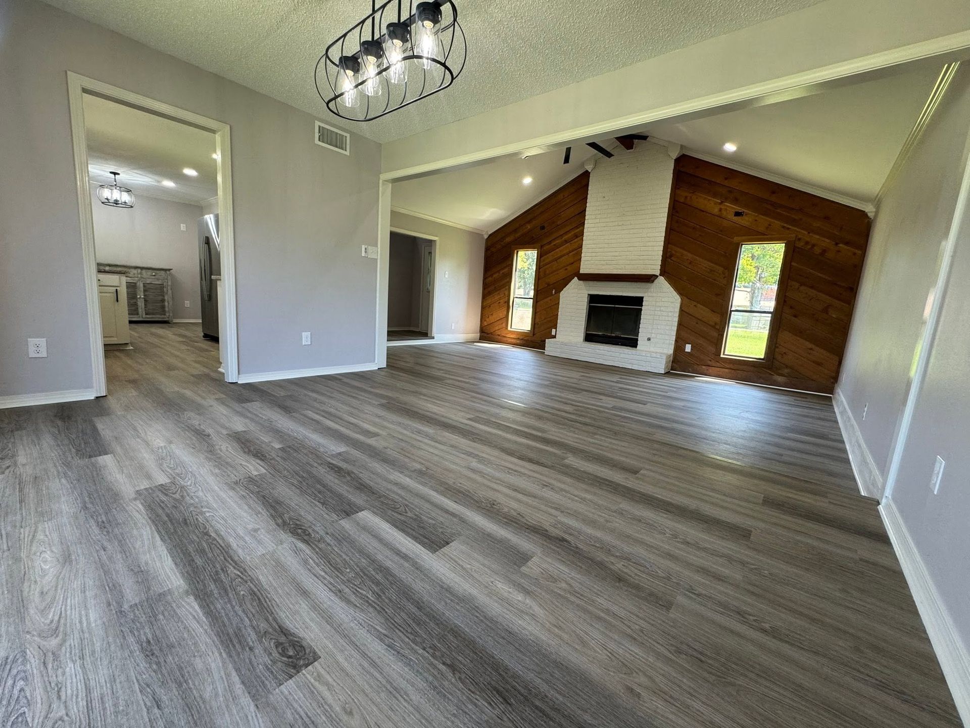Empty open living room with gray wood floors, vaulted ceiling, fireplace, and brick accent wall