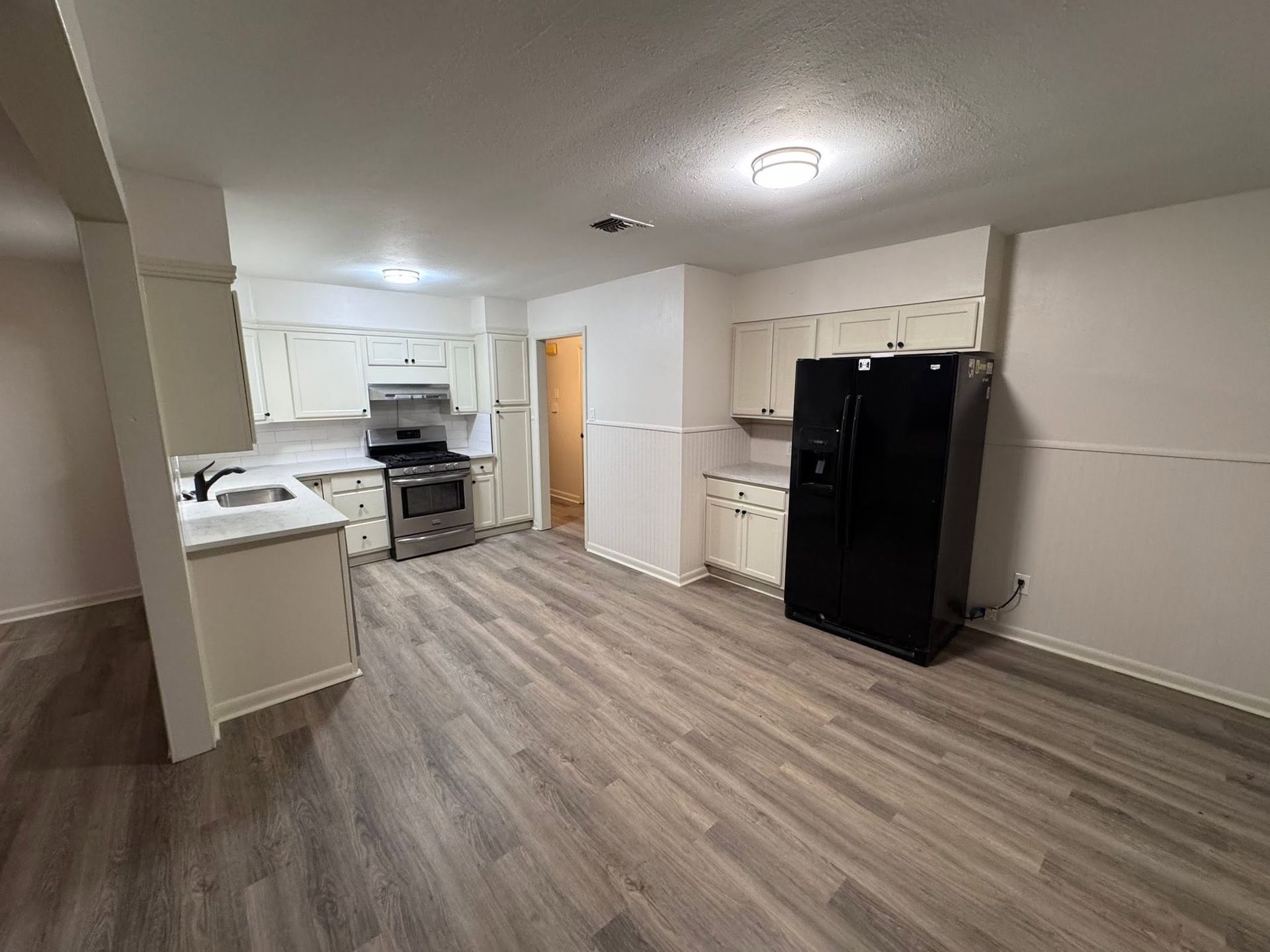 Empty kitchen with white cabinets, wood-look flooring, black refrigerator, and stainless steel stove.