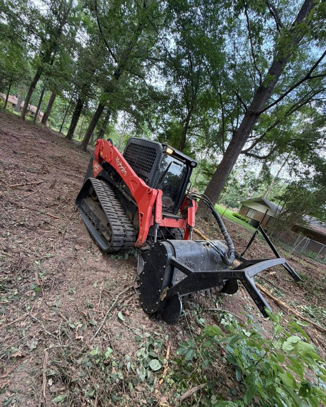 Orange skid steer with grapple attachment on a wooded dirt slope.