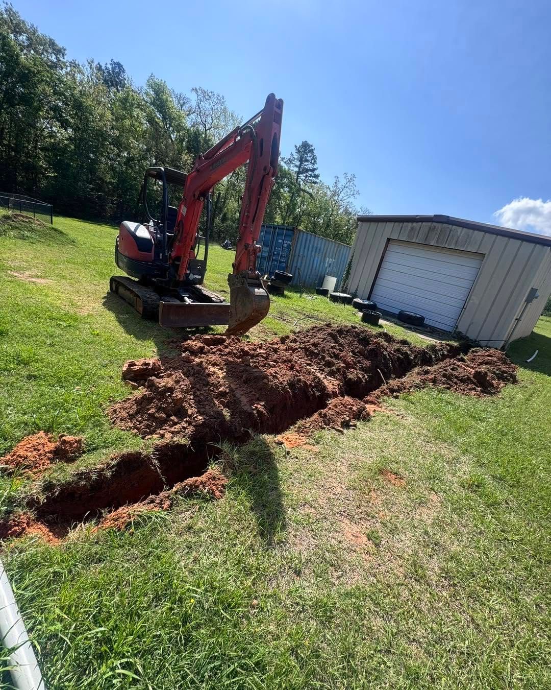 Red mini excavator digging a trench beside a white shed on a sunny grassy lot.