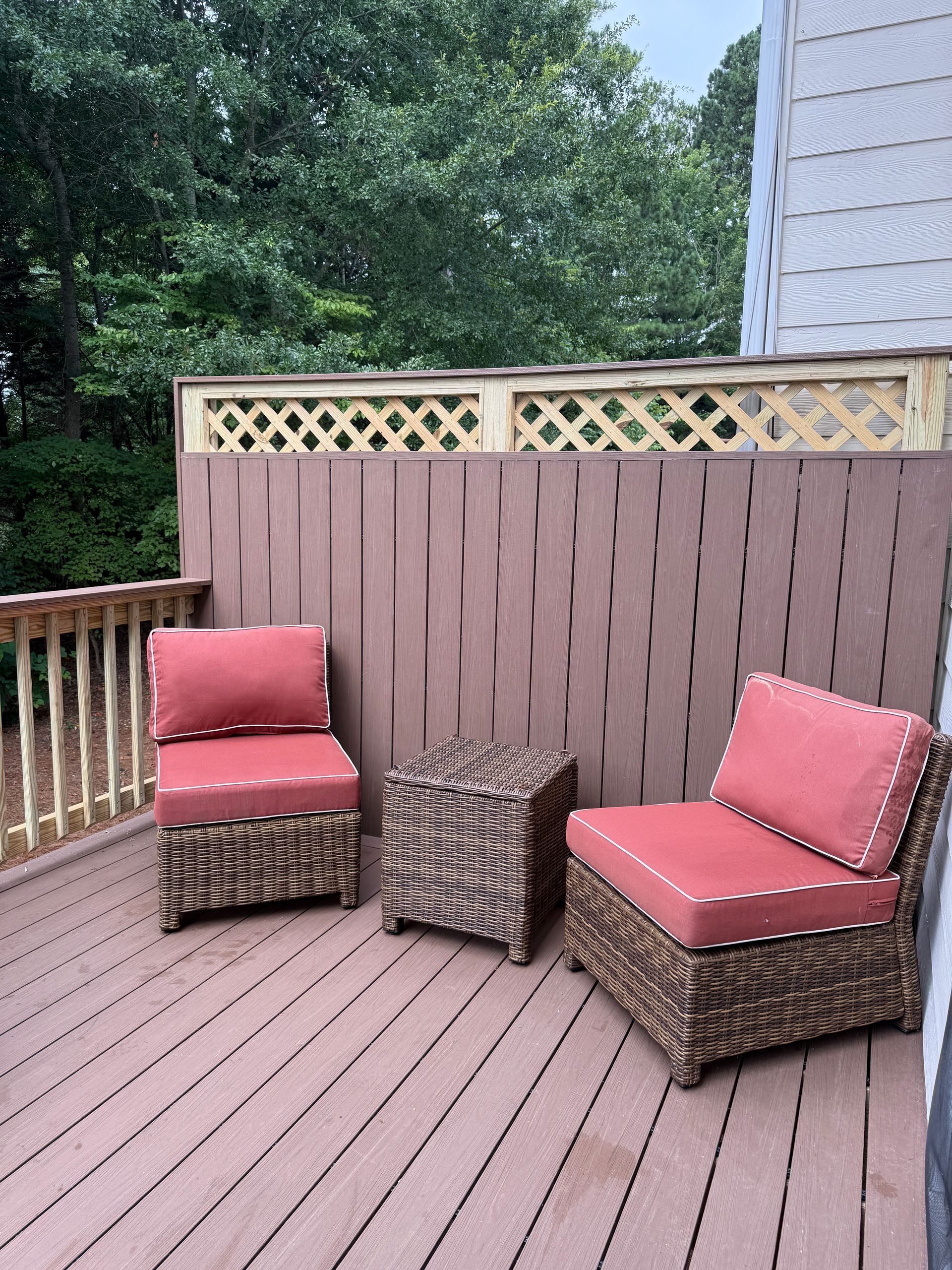 Outdoor deck with two red cushioned chairs and a small table, surrounded by wood railing and trees.