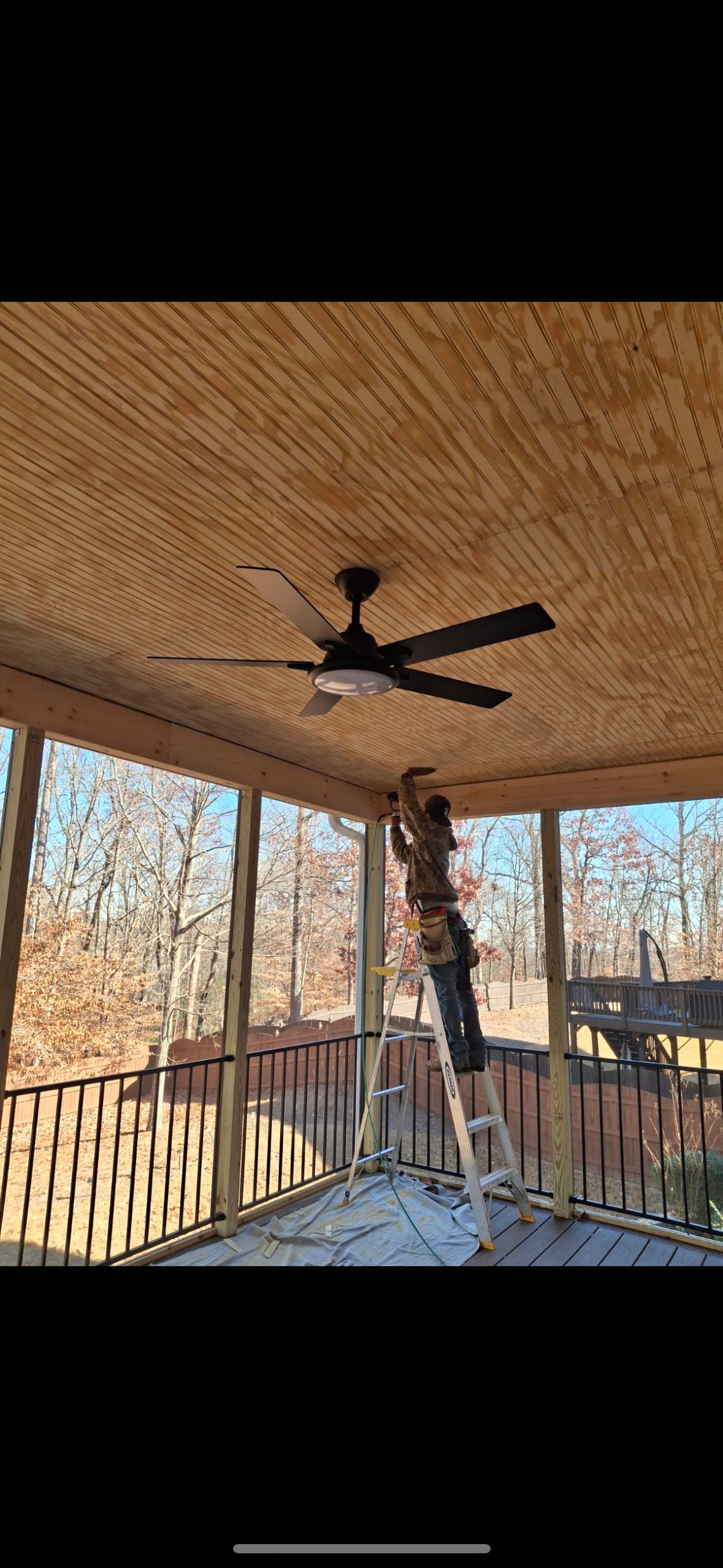 Covered porch with a ceiling fan, railing, and a person on a ladder near hanging decorations