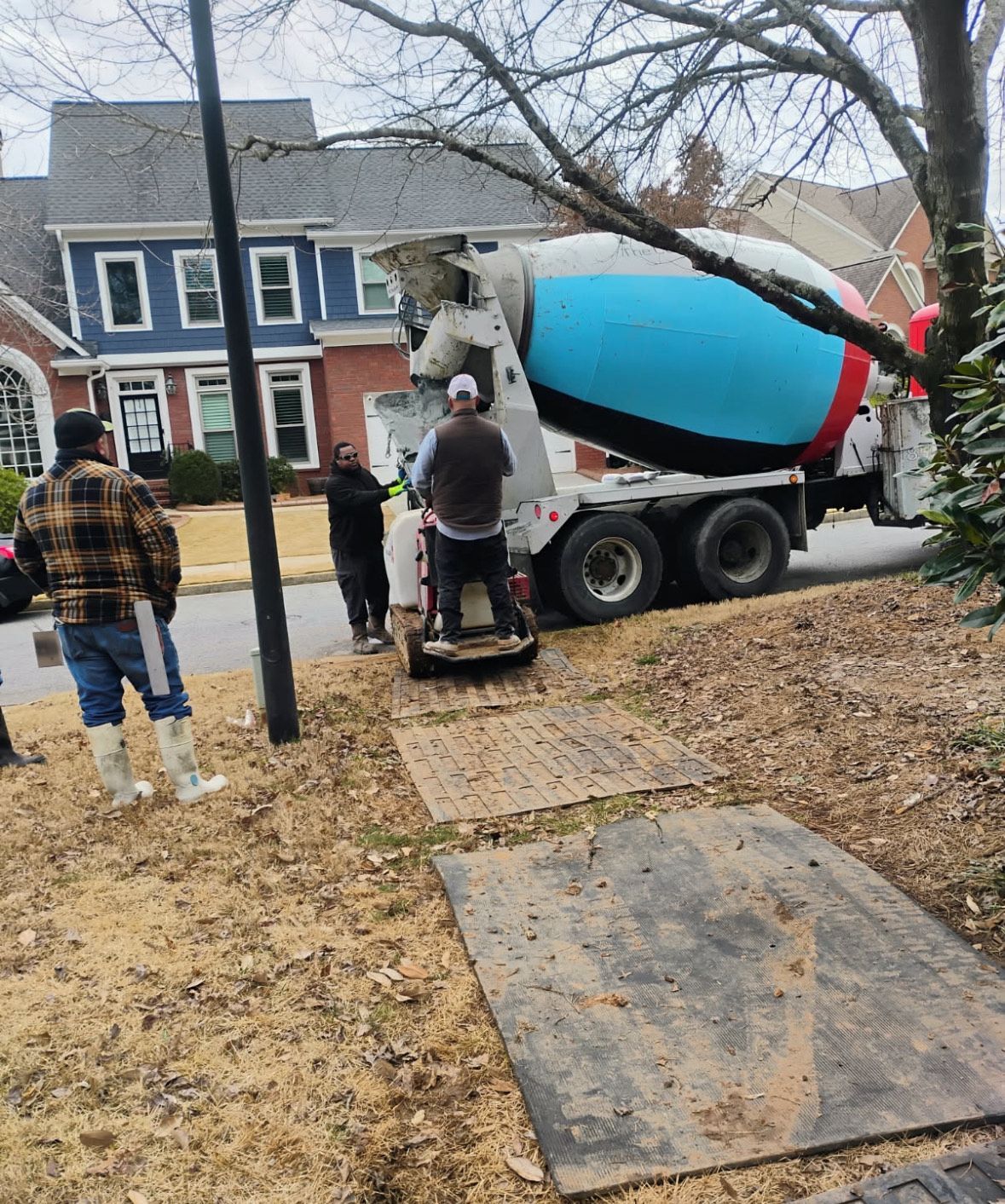 Concrete truck pouring driveway near houses; two workers stand nearby in a suburban yard.