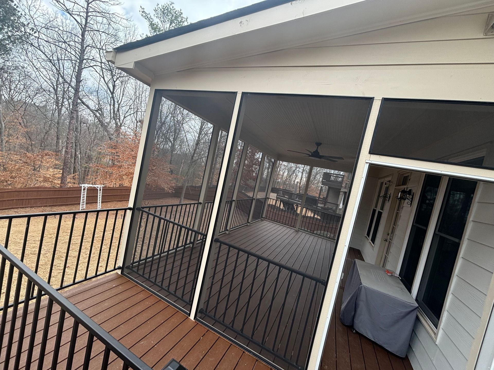 Screened porch with brown deck, black railing, and gray-covered grill overlooking trees