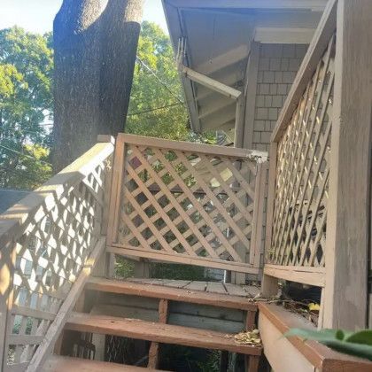 Wooden outdoor stairs and lattice railing beside a house, with a large tree and green foliage nearby.