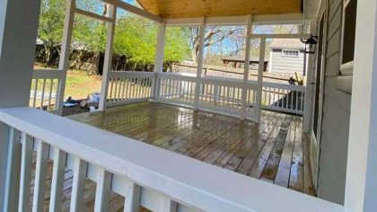 Covered wooden porch with white railings and posts, overlooking a yard on a sunny day.