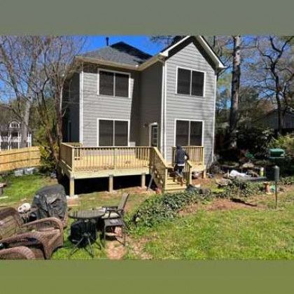 Gray two-story house with a wooden deck and yard, surrounded by trees under a clear blue sky